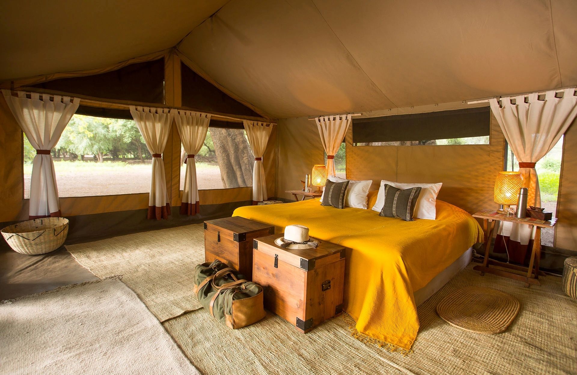 A spacious safari tent interior with a bed featuring a bright yellow throw, wooden trunks at the foot, and canvas walls.