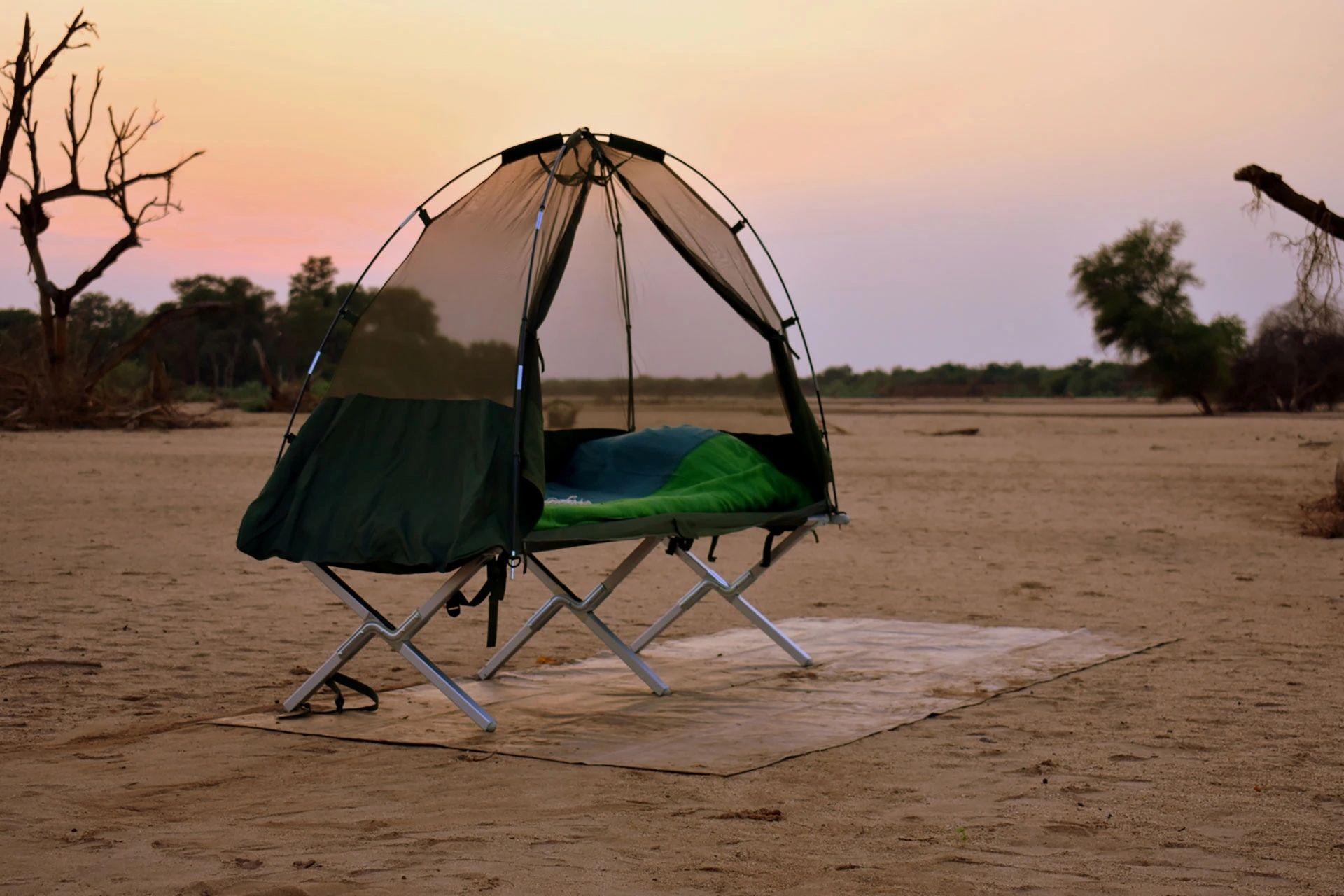 A portable tent cot with a mesh top and green sleeping bag set up on a mat in a sandy, desolate landscape at sunset.