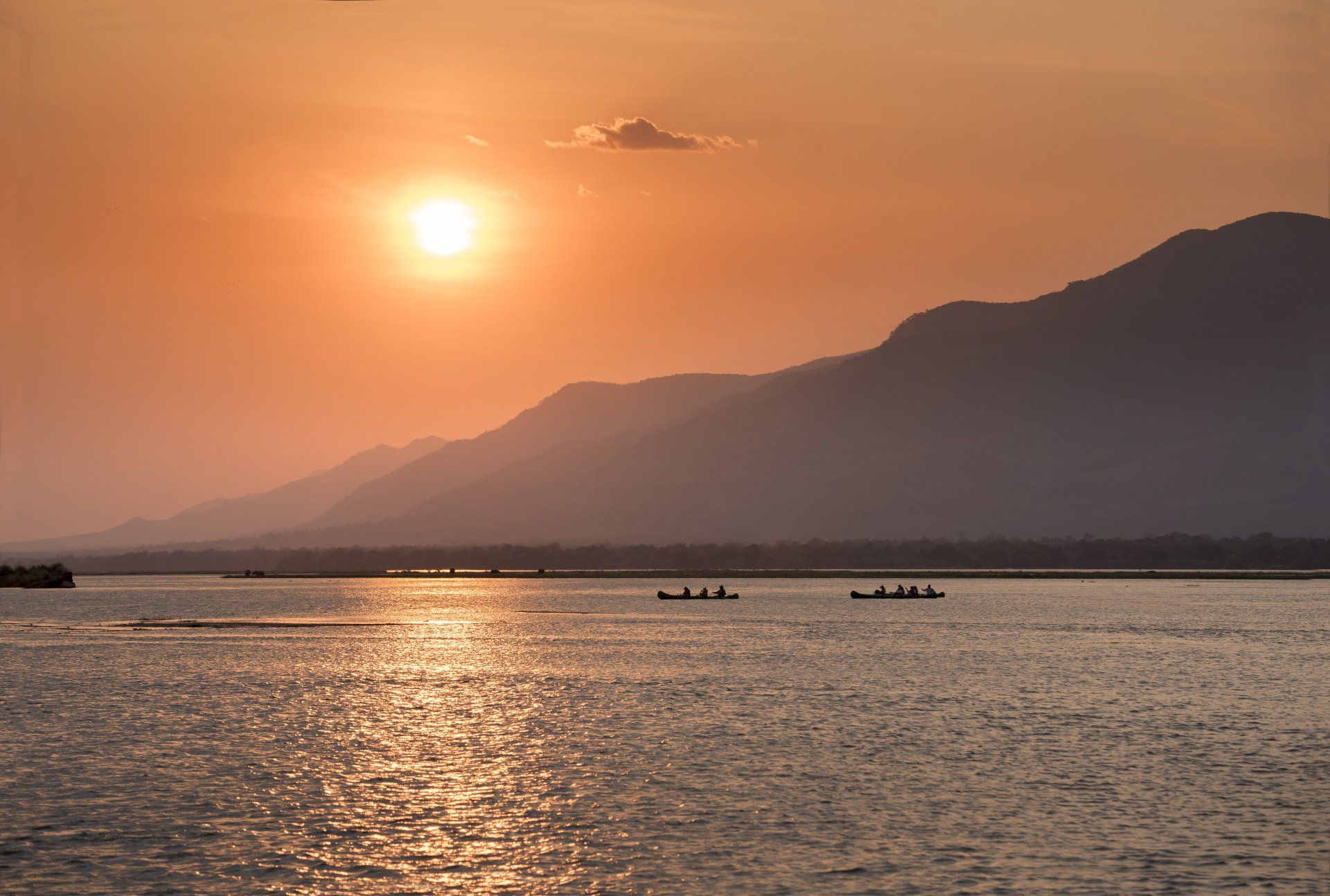 Sun setting over a mountain range and a calm lake with two small boats in the distance.