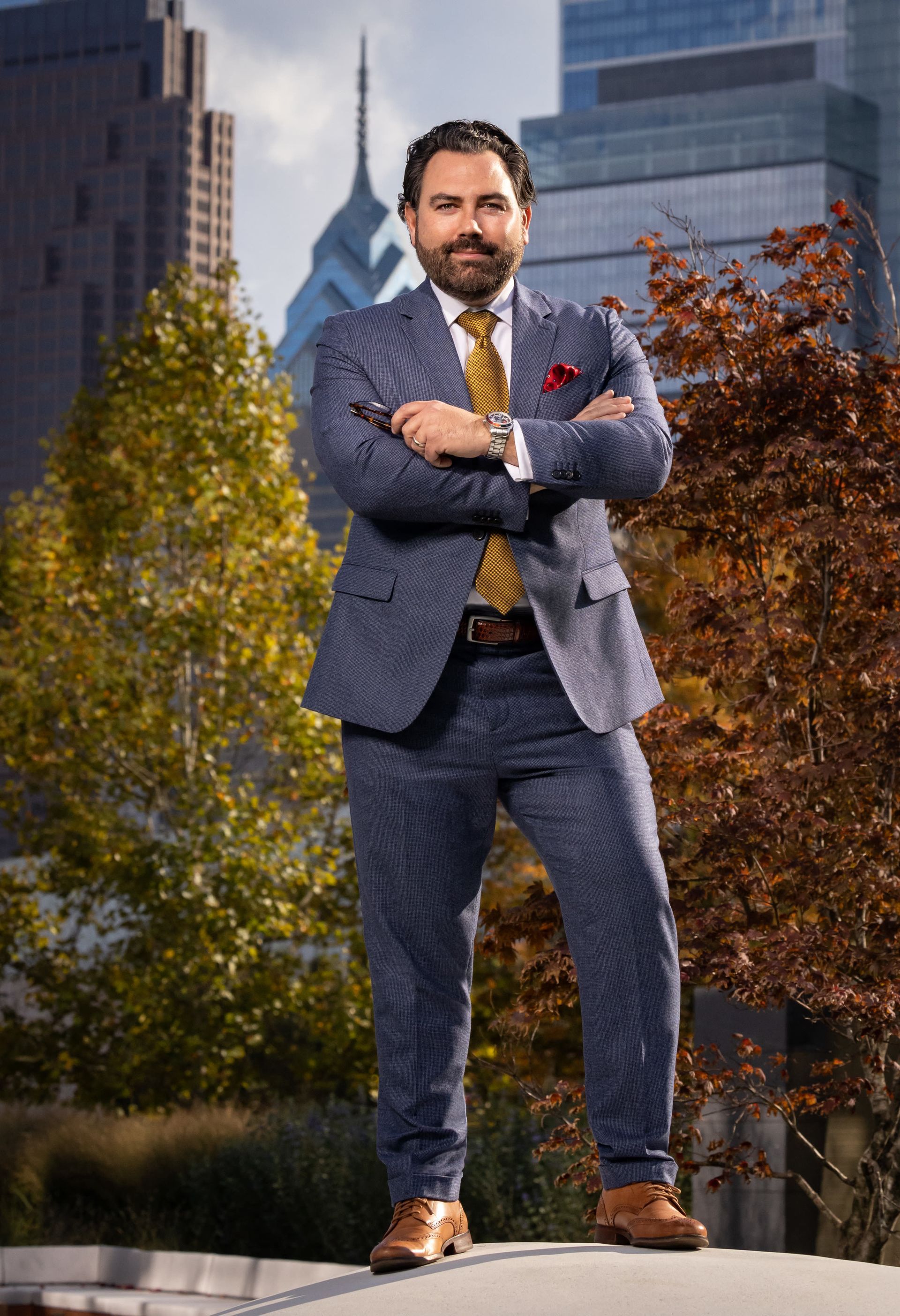 Man in suit with arms crossed, in front of Philadelphia skyline.