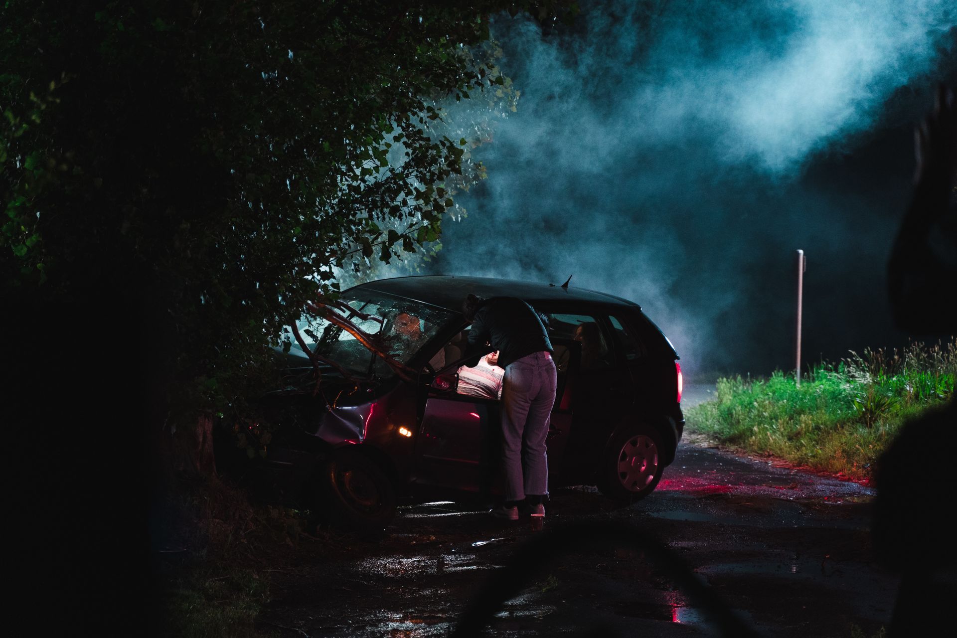 Car crashed at night, person standing beside it, smoke and dim lighting present.