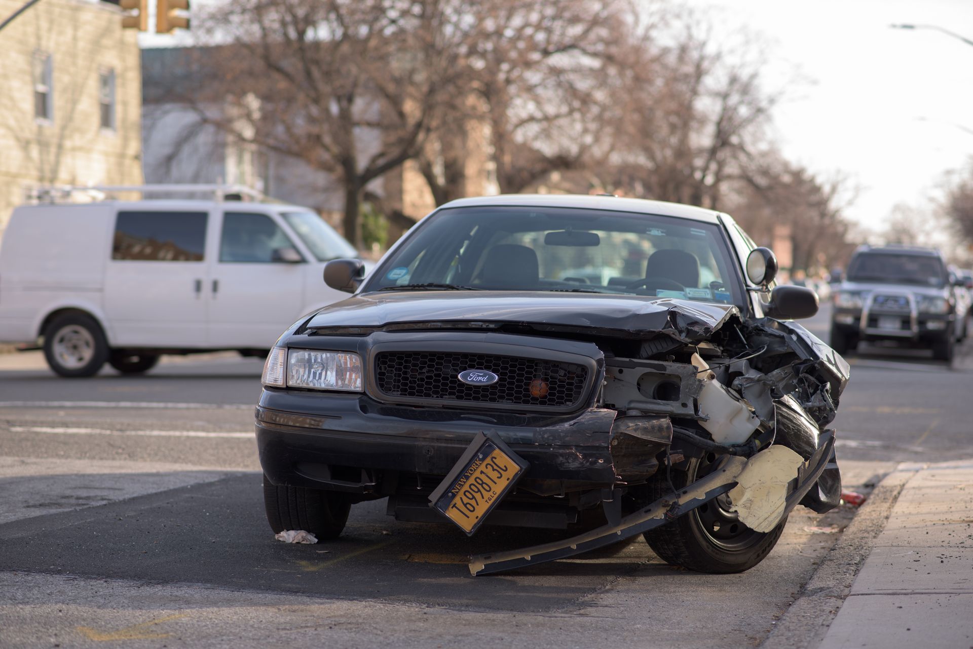 Black car with extensive front-end damage parked on a street; white van and other vehicles in the background.