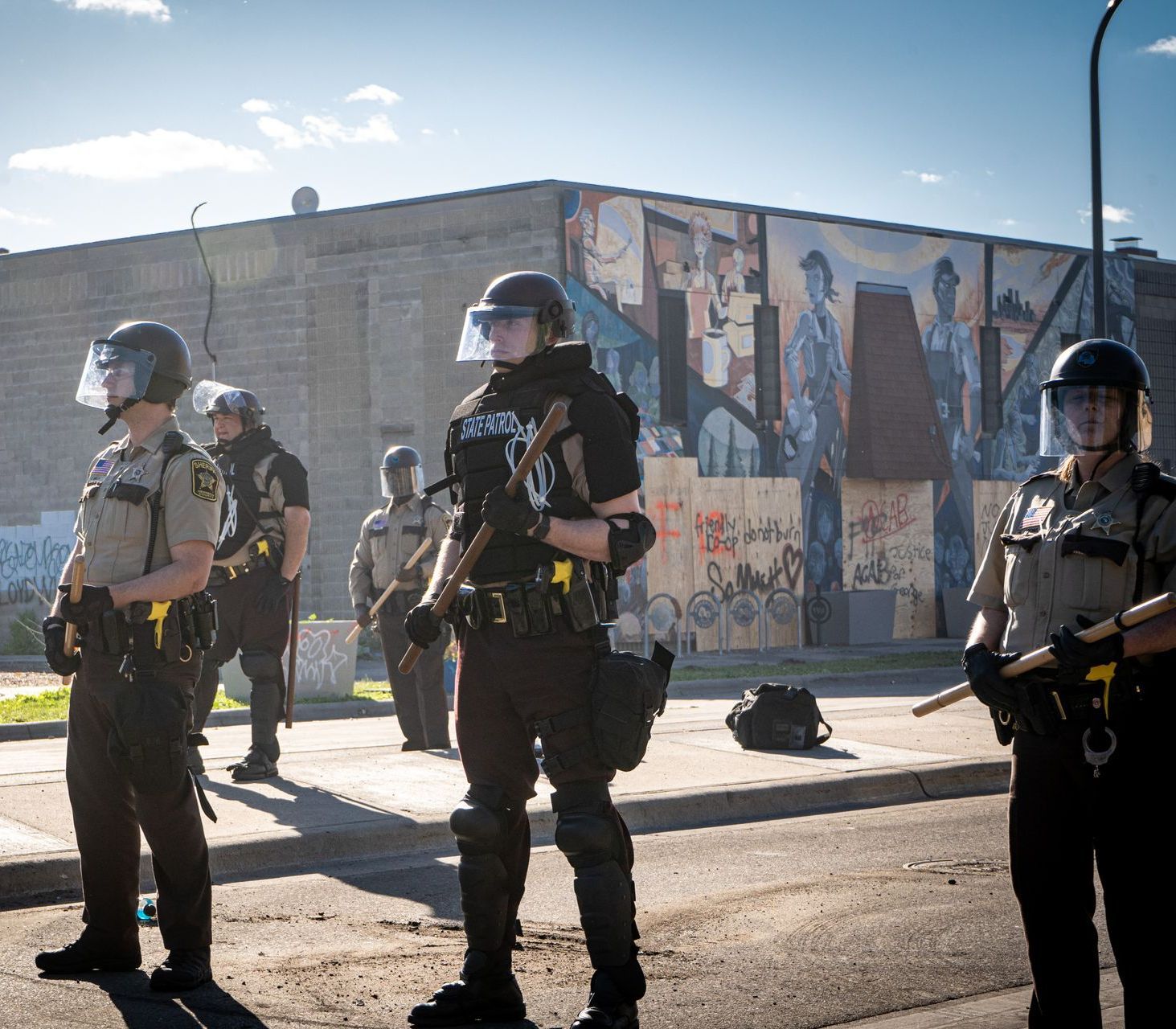 Police officers in riot gear stand on a street, facing a building with a mural in the background.