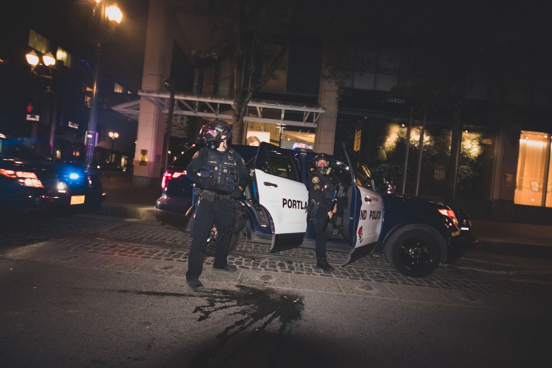 Police officers in riot gear stand near a Portland police SUV on a city street at night.