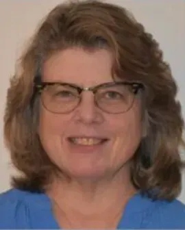 Smiling person with glasses and shoulder-length brown hair against a plain background