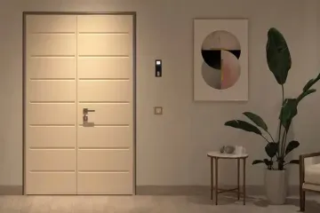 Ingresso moderno con porta beige illuminata, quadro astratto, tavolino e pianta alta in vaso.
