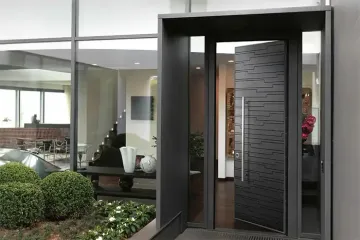 Ingresso principale moderno con porta scura dalla texture particolare, accanto a grandi finestre adibite a ufficio e al paesaggio circostante.