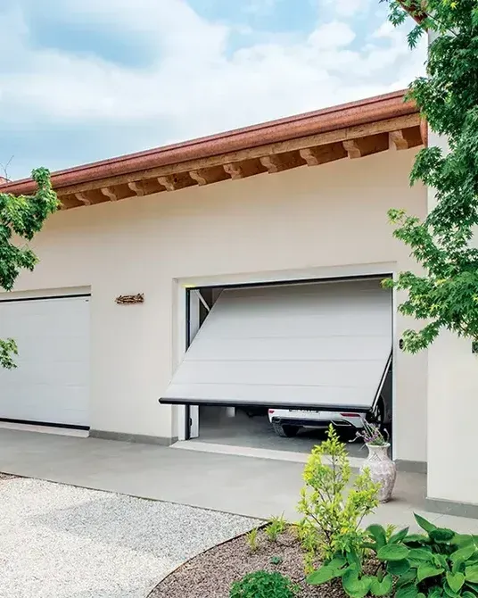 La porta del garage è socchiusa su una casa beige, con un'auto bianca all'interno e alberi che incorniciano il vialetto d'accesso.