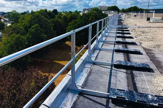 Passerella sul tetto con ringhiera metallica e pannelli neri sparsi, affacciata sugli alberi sotto un cielo azzurro.