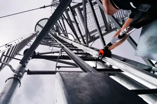 Un operaio si arrampica su una torre metallica con corde e dispositivi di sicurezza, sullo sfondo di un cielo grigio.
