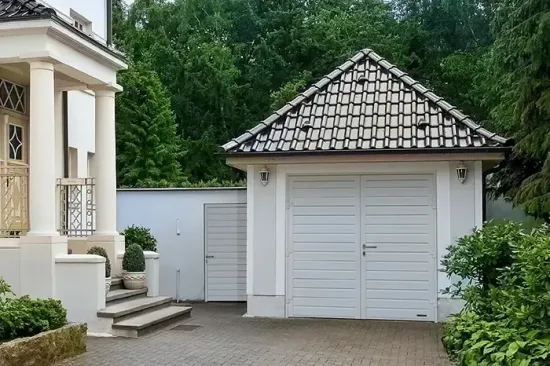 Garage bianco con tetto in tegole accanto a una casa color crema e a una rigogliosa vegetazione in un vialetto pavimentato.