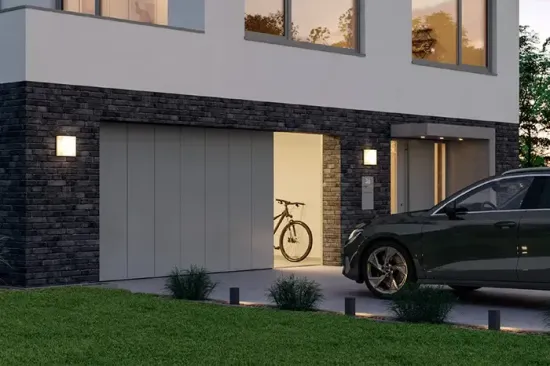 Esterno di una casa moderna al crepuscolo, con garage illuminato, auto parcheggiata e bicicletta visibili all'interno.