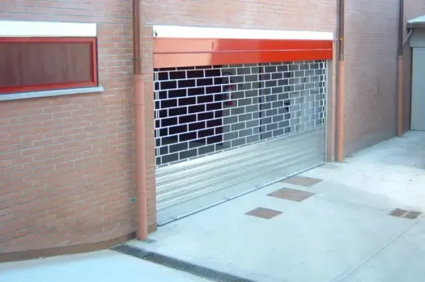 Serranda di sicurezza metallica chiusa all'ingresso di un edificio in mattoni con finiture rosse.