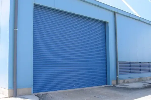 Porta avvolgibile industriale blu su un edificio adibito a magazzino di colore azzurro chiaro.