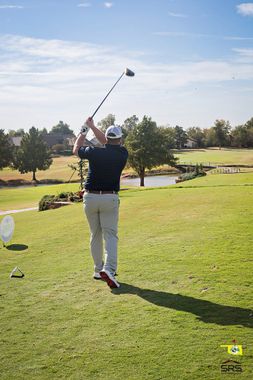 Golfer swinging a club on a sunny golf course; green grass, water, trees in the background.