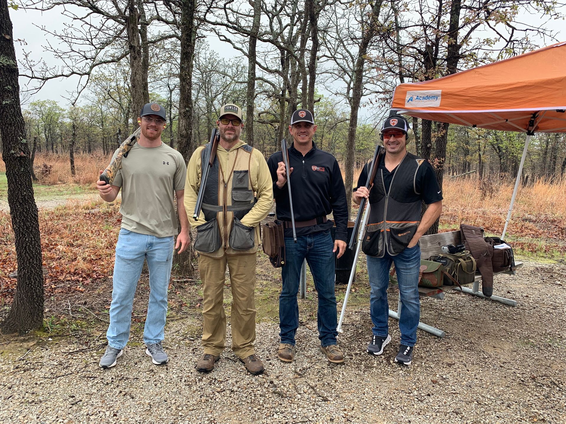 Four men stand with shotguns in a wooded area, posing near an orange tent and shooting gear.