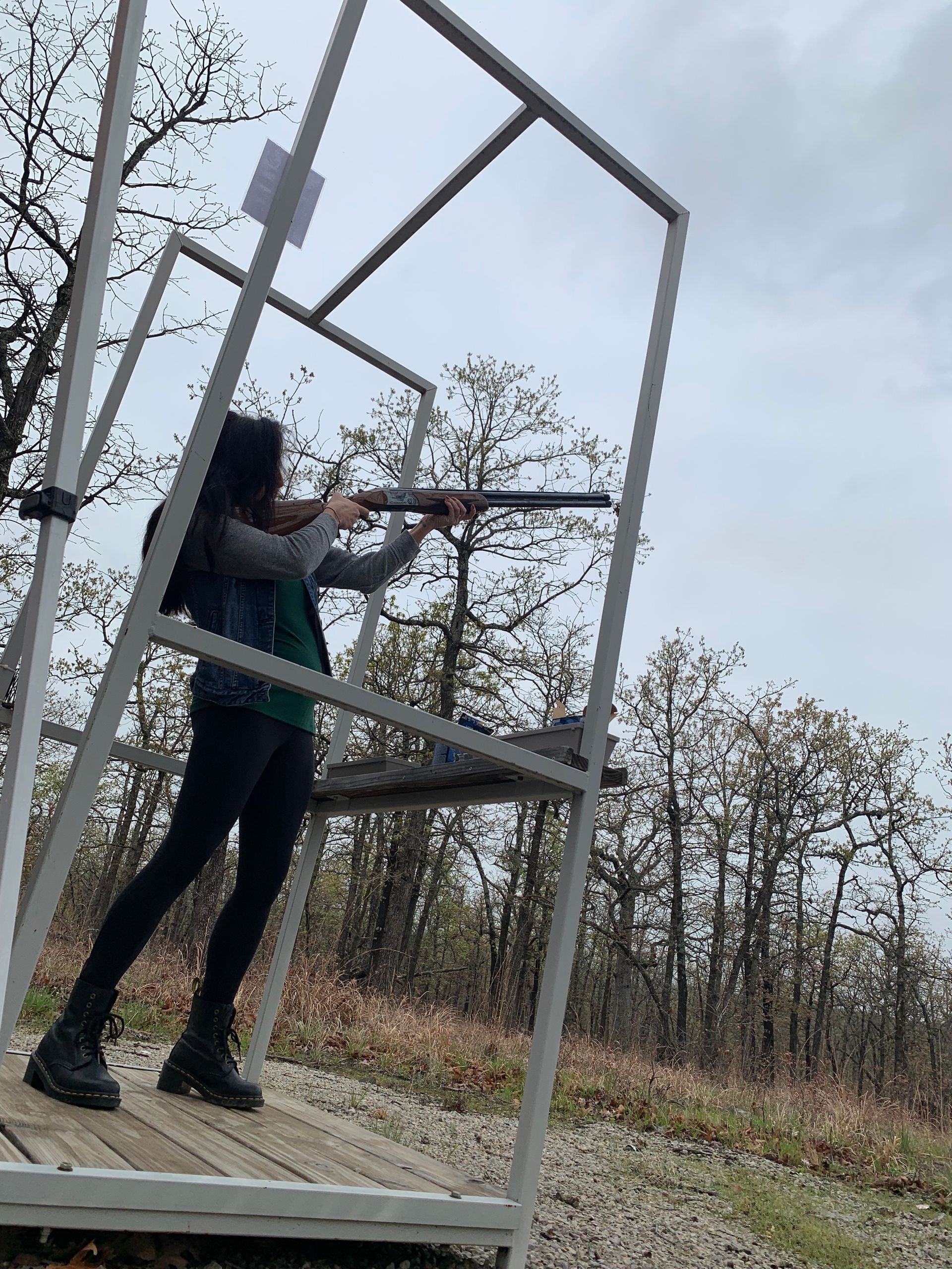 Woman shooting a shotgun at a skeet range. She stands inside a metal frame, aiming at the sky.