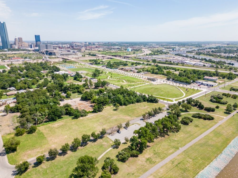 Aerial view of a park with trees, pathways, and green space, with a city skyline in the background.