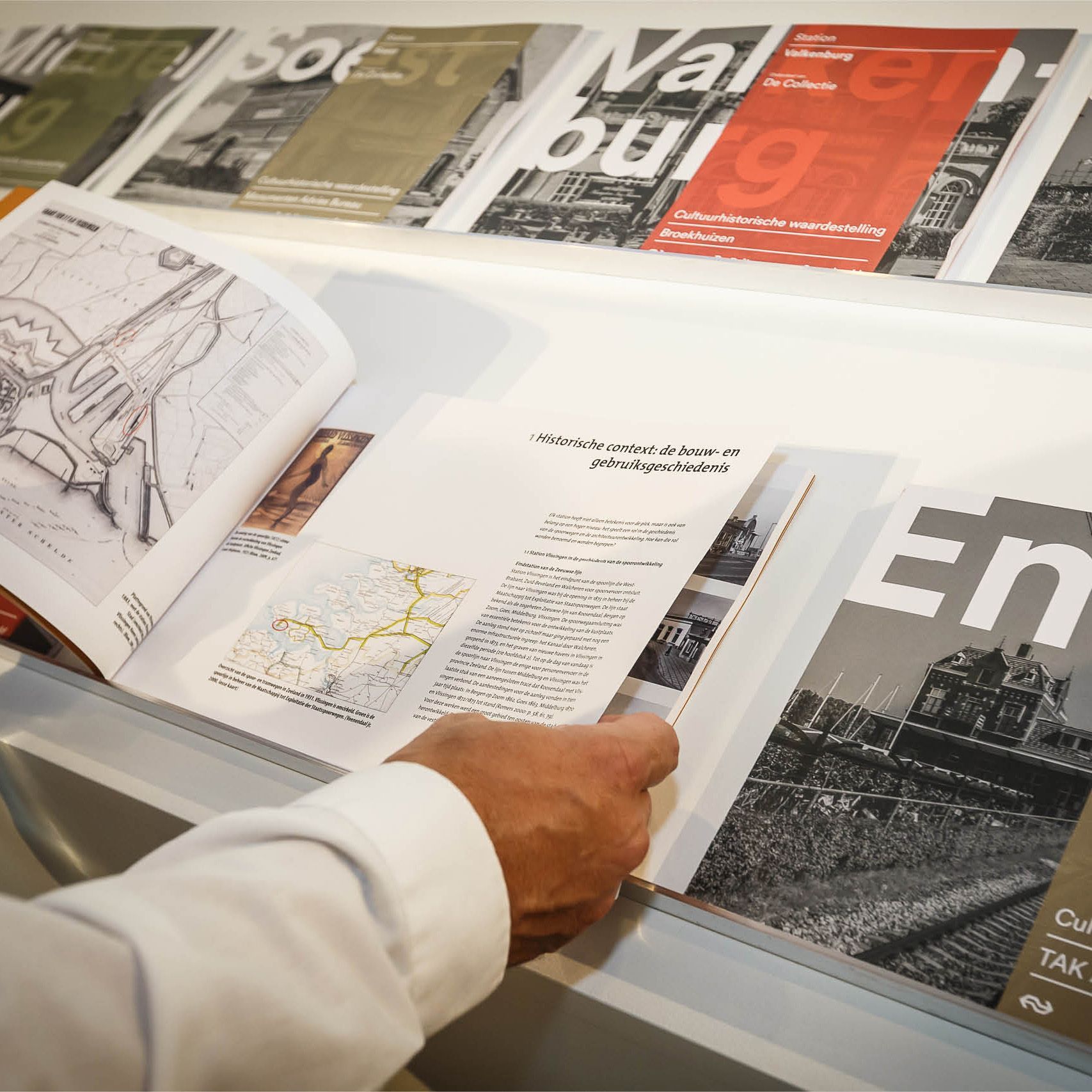 Hand bladert door tijdschriften op een displayplank, met architectonische ontwerpen en tekst.