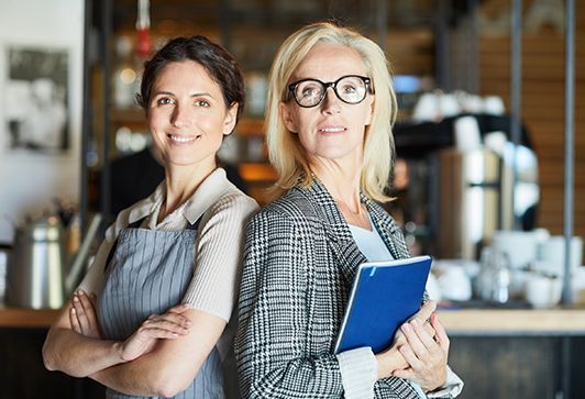 Two women in a cafe: one in apron, the other in blazer, both smiling, arms crossed, one holding a blue notebook.