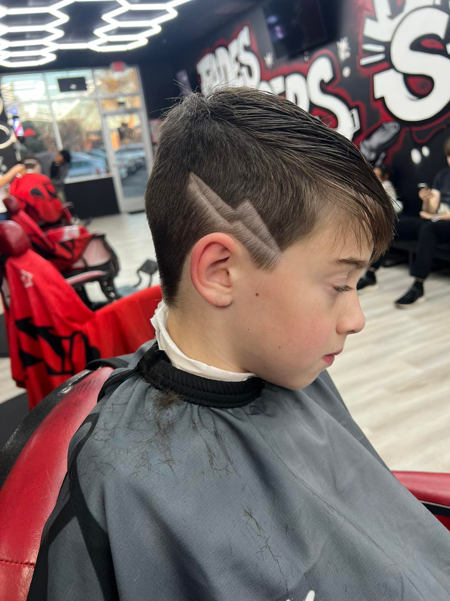 Child with a fresh fade haircut in a barber chair, wearing a black cape in a barbershop.