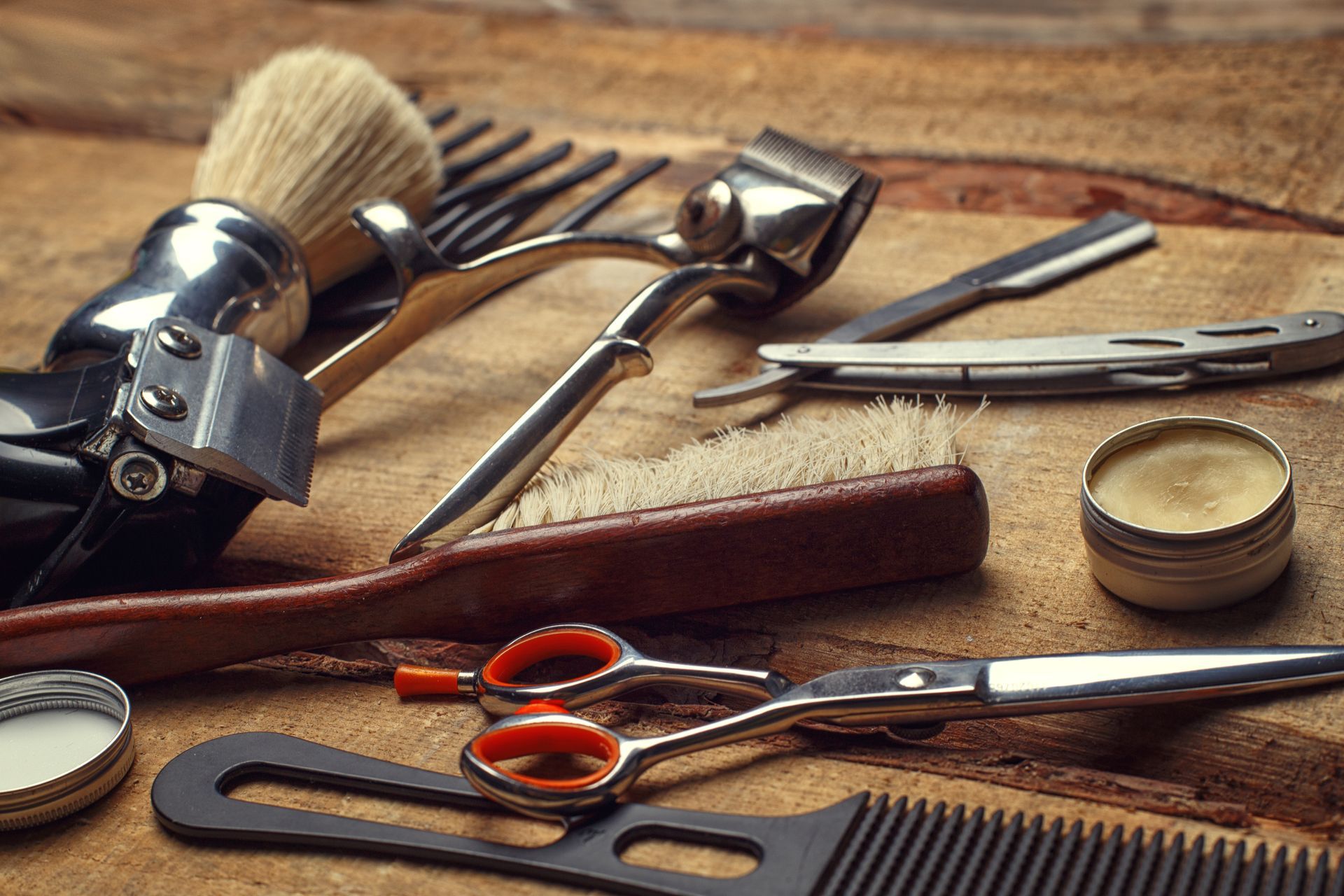 Assorted barbering tools on a wooden table, including scissors, combs, brush, razor, and grooming accessories.