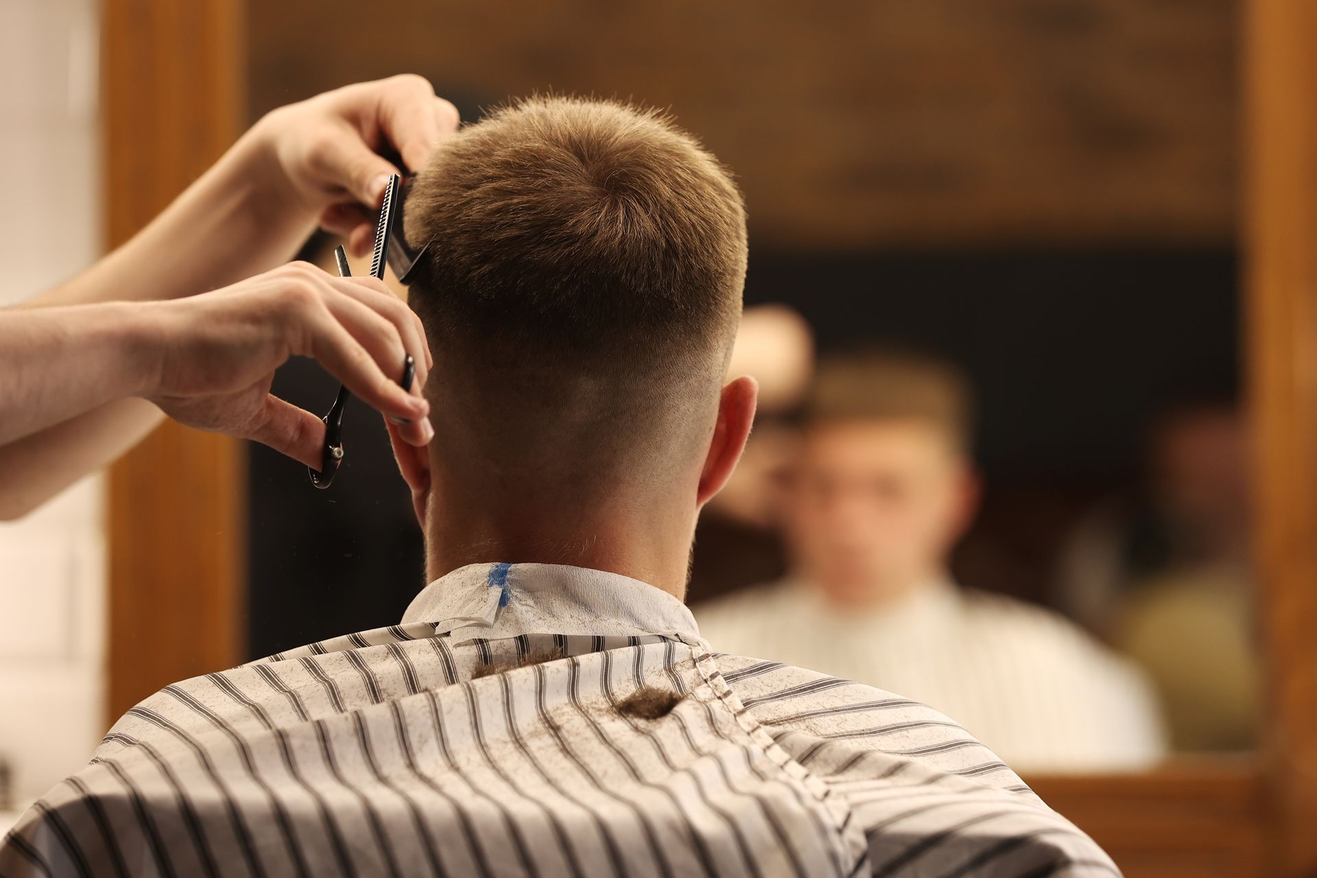 Barber trimming a customer’s hair in front of a mirror, with striped cape in a warm-lit barbershop