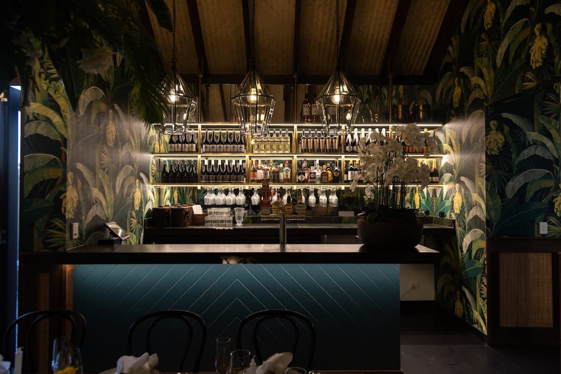 A bar in a restaurant with a tropical wallpaper on the wall.