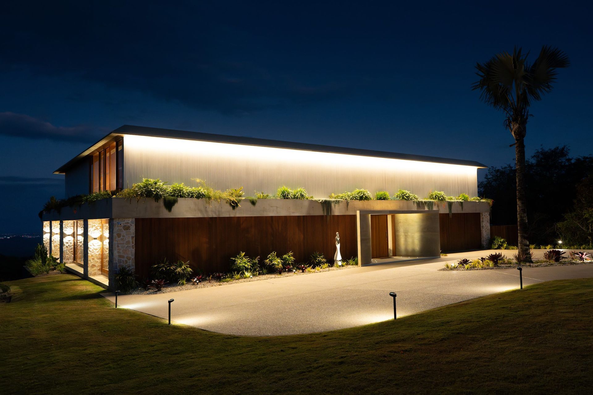 Modern house at night, illuminated with strip lighting, driveway lit by small ground lights, dark blue sky.