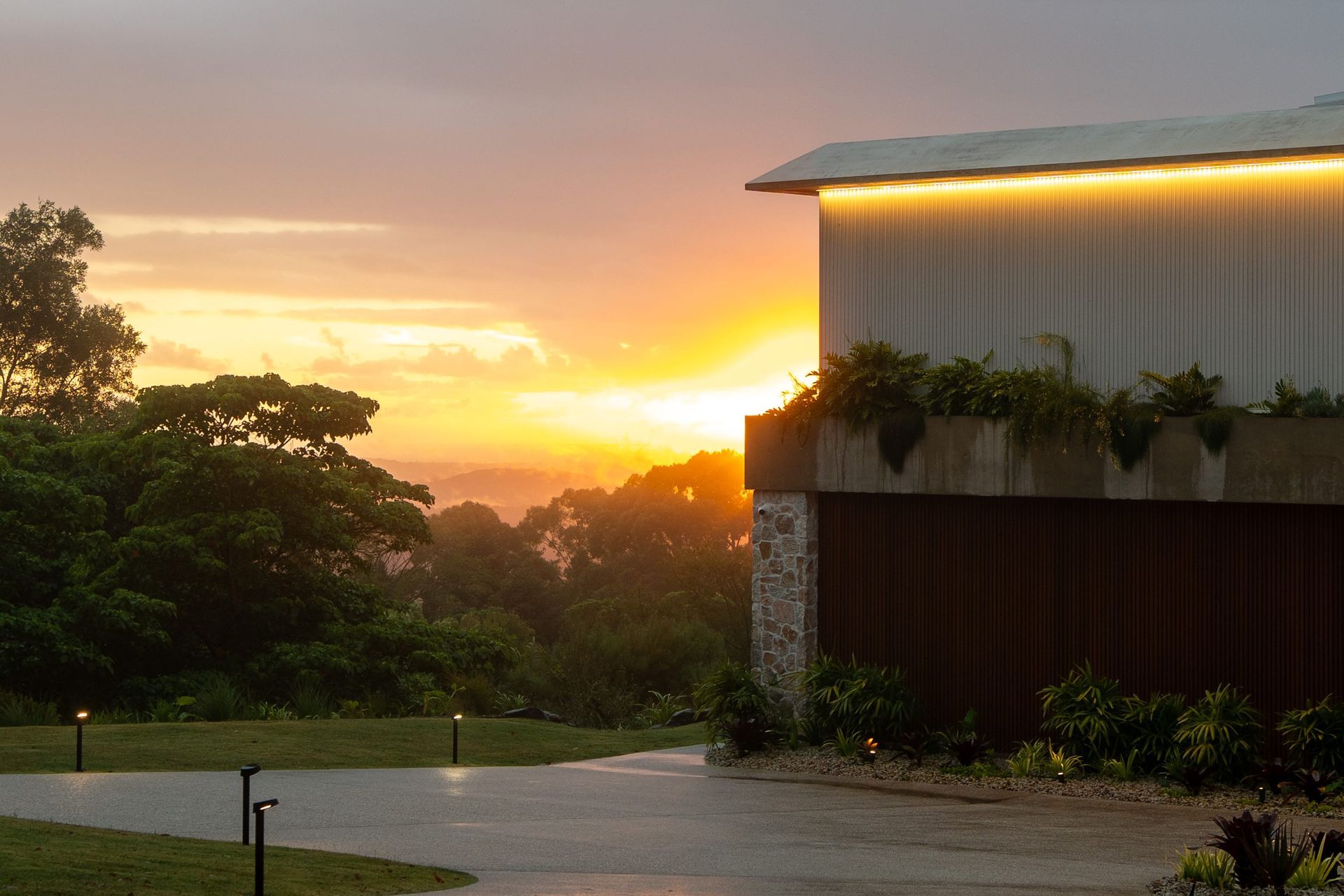 Sunset over building with a green roof, lush trees, and a wet driveway.