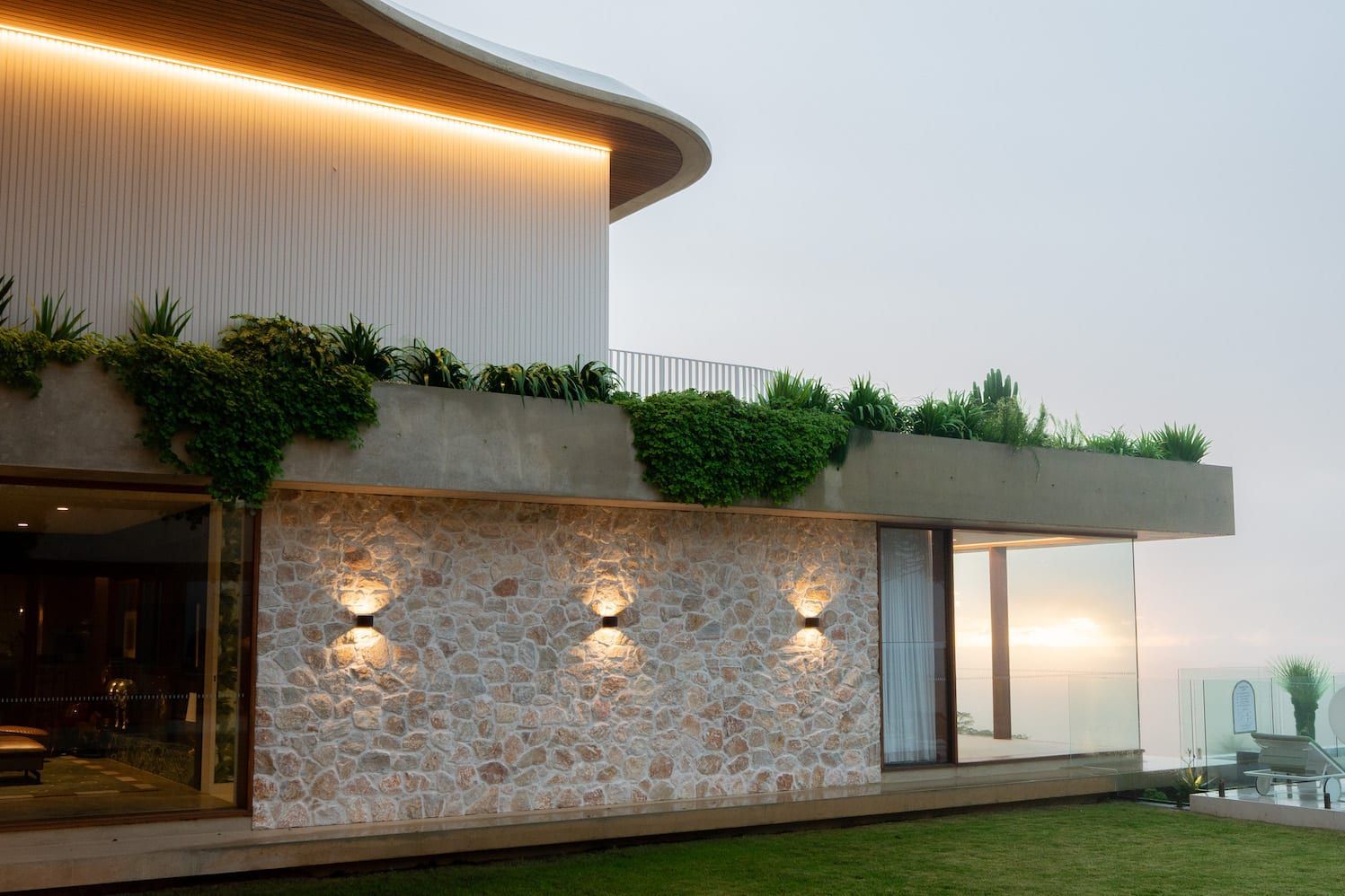Modern house exterior with stone wall, greenery, and outdoor lighting.