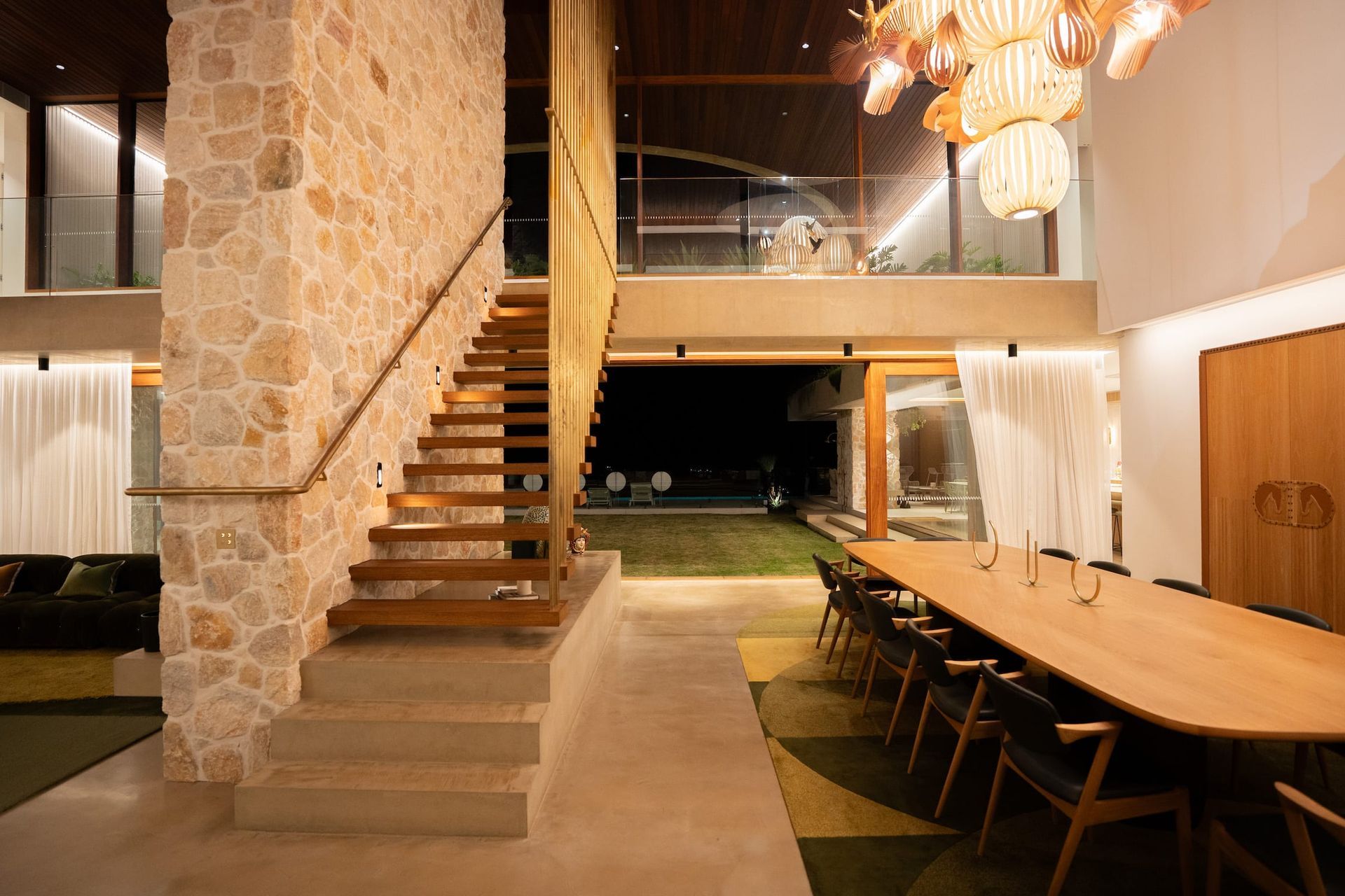 Interior with stone wall, wooden stairs, large dining table, and view to a backyard at night.