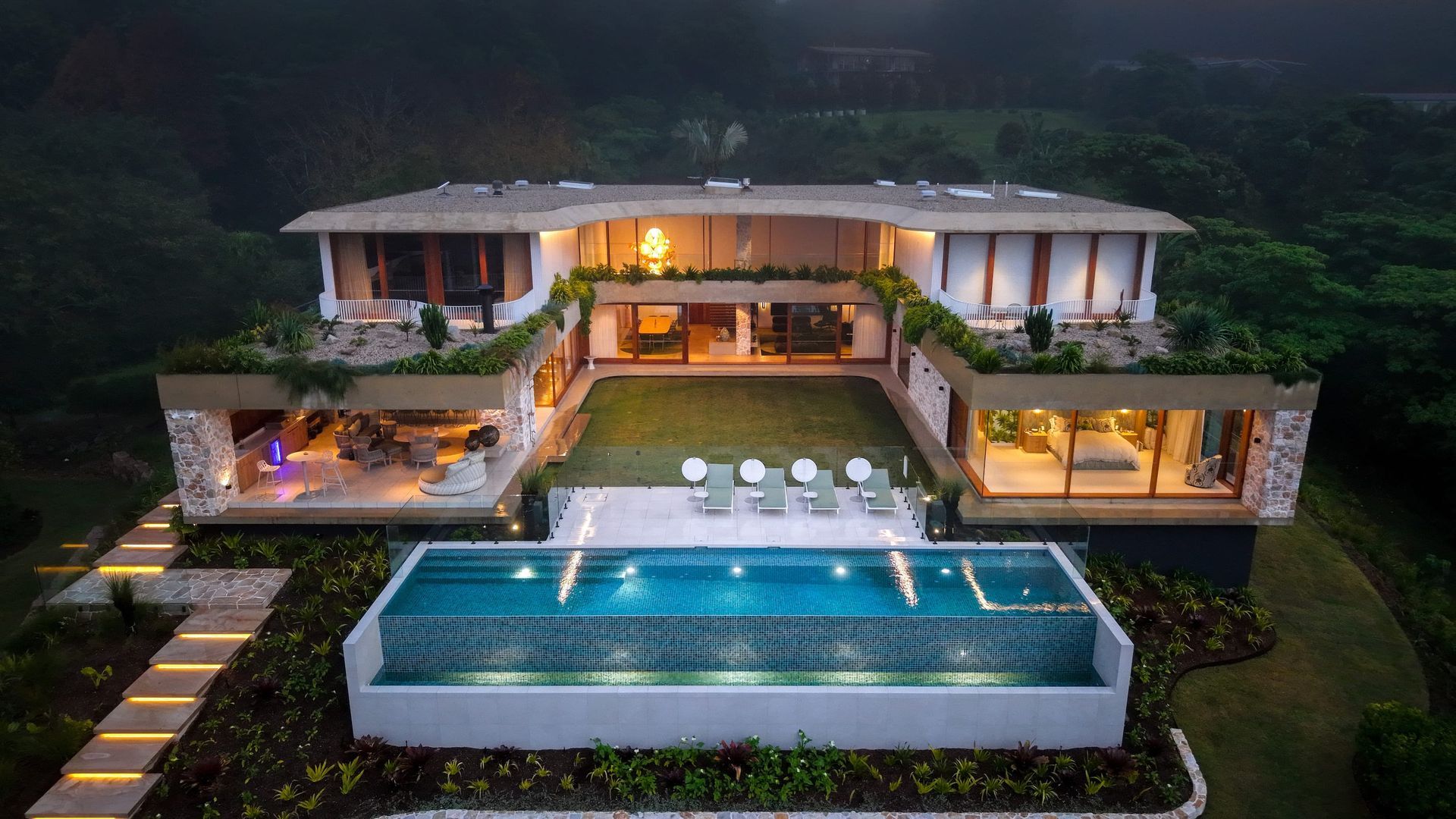Luxury home with pool overlooking a hillside. Blue pool, stone patio, green grass, and modern architecture.