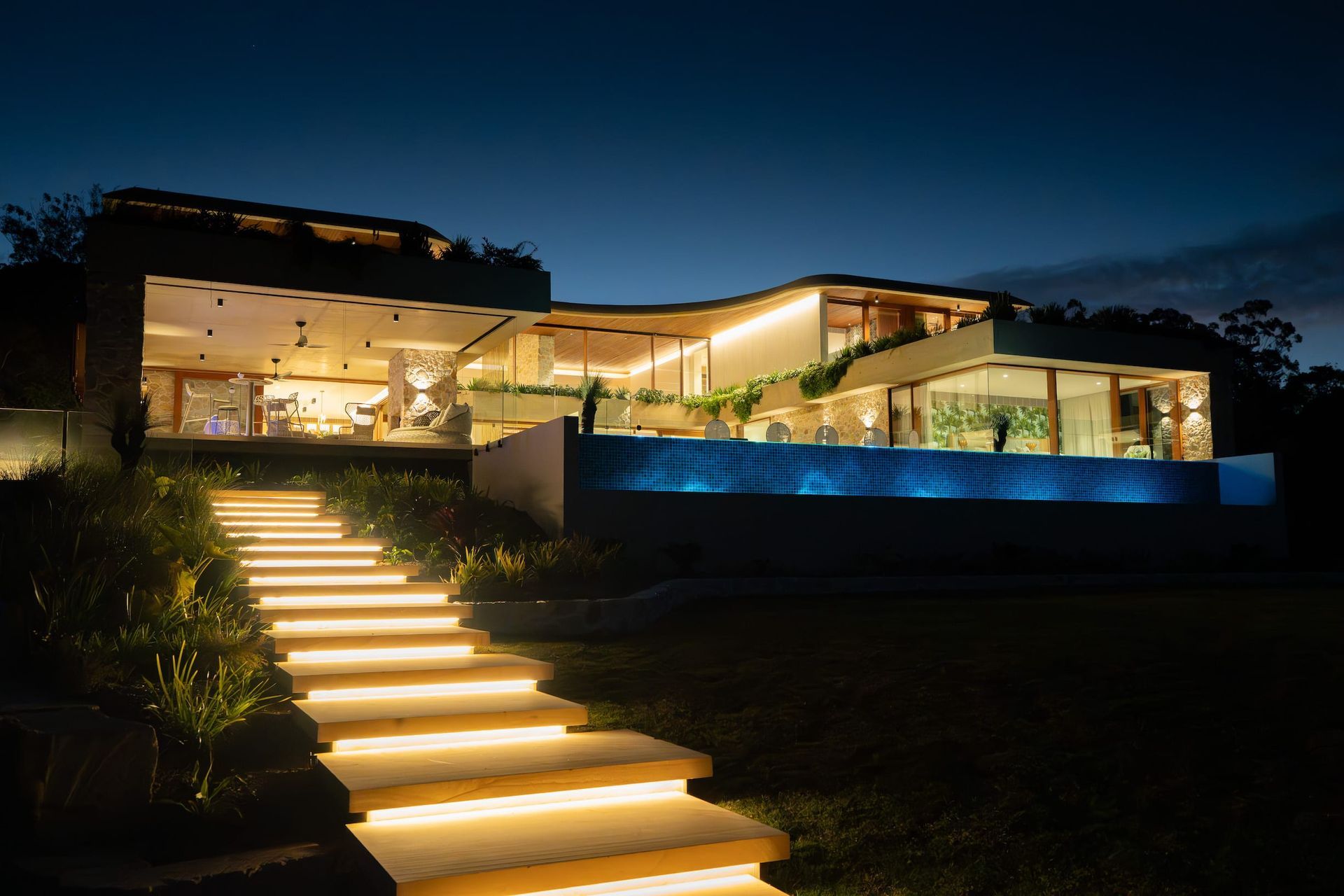 Lit staircase leads to a modern home with glowing lights and a pool at night.
