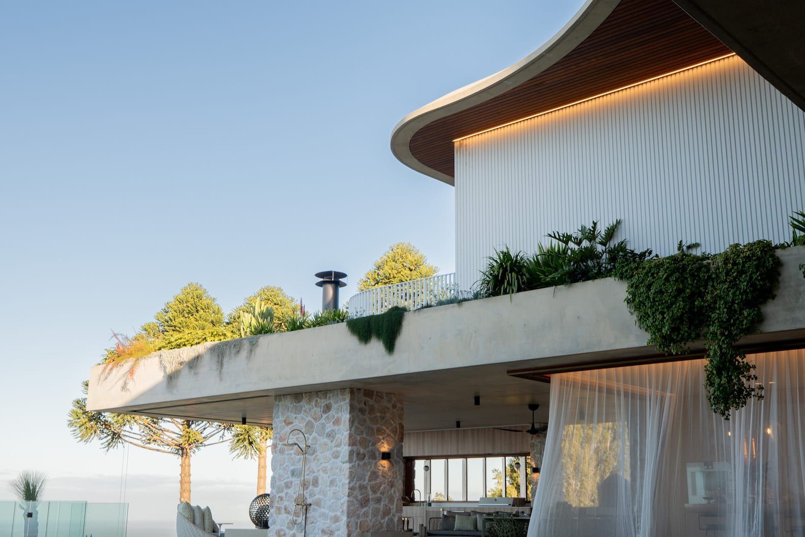 A modern building with a curved roof, greenery, and stone accents, with a clear sky in the background.