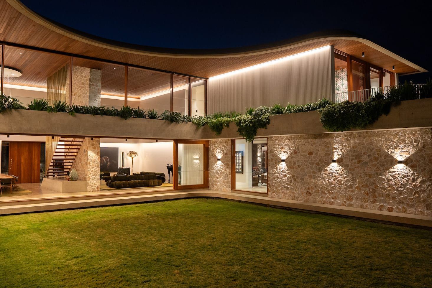 Modern home exterior at night with stone walls, curved roof, and illuminated lawn.