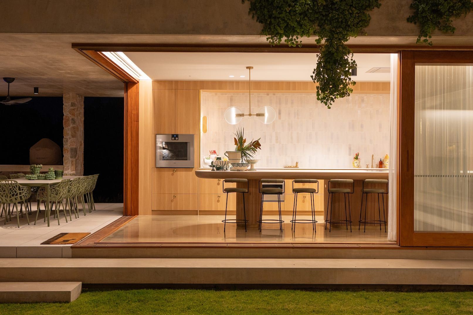 Outdoor kitchen with open sliding door. Wooden cabinetry and countertop, overlooking a dining area with a green lawn.