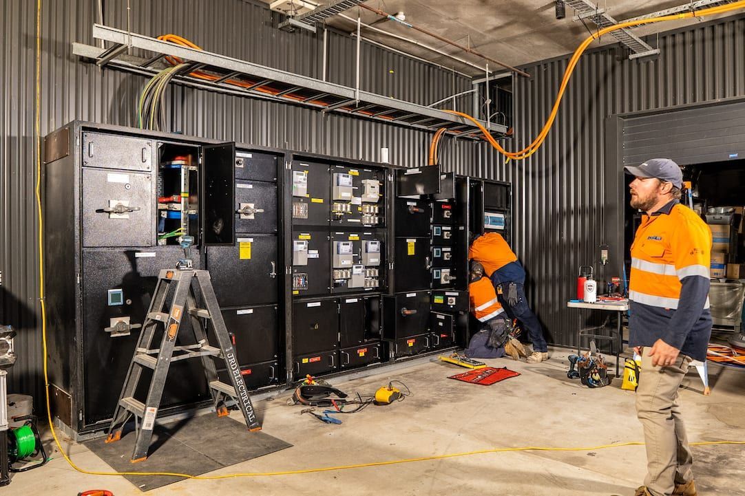 Two workers in orange safety gear inspect electrical cabinets in an industrial room.