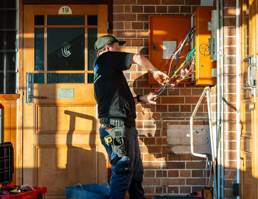 Worker in black shirt installing wiring on a brick house porch beside a wooden door and orange utility box