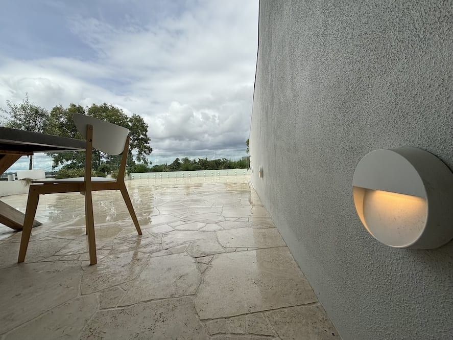 Patio with stone floor, wooden chair, and round wall light overlooking a cloudy beachside view
