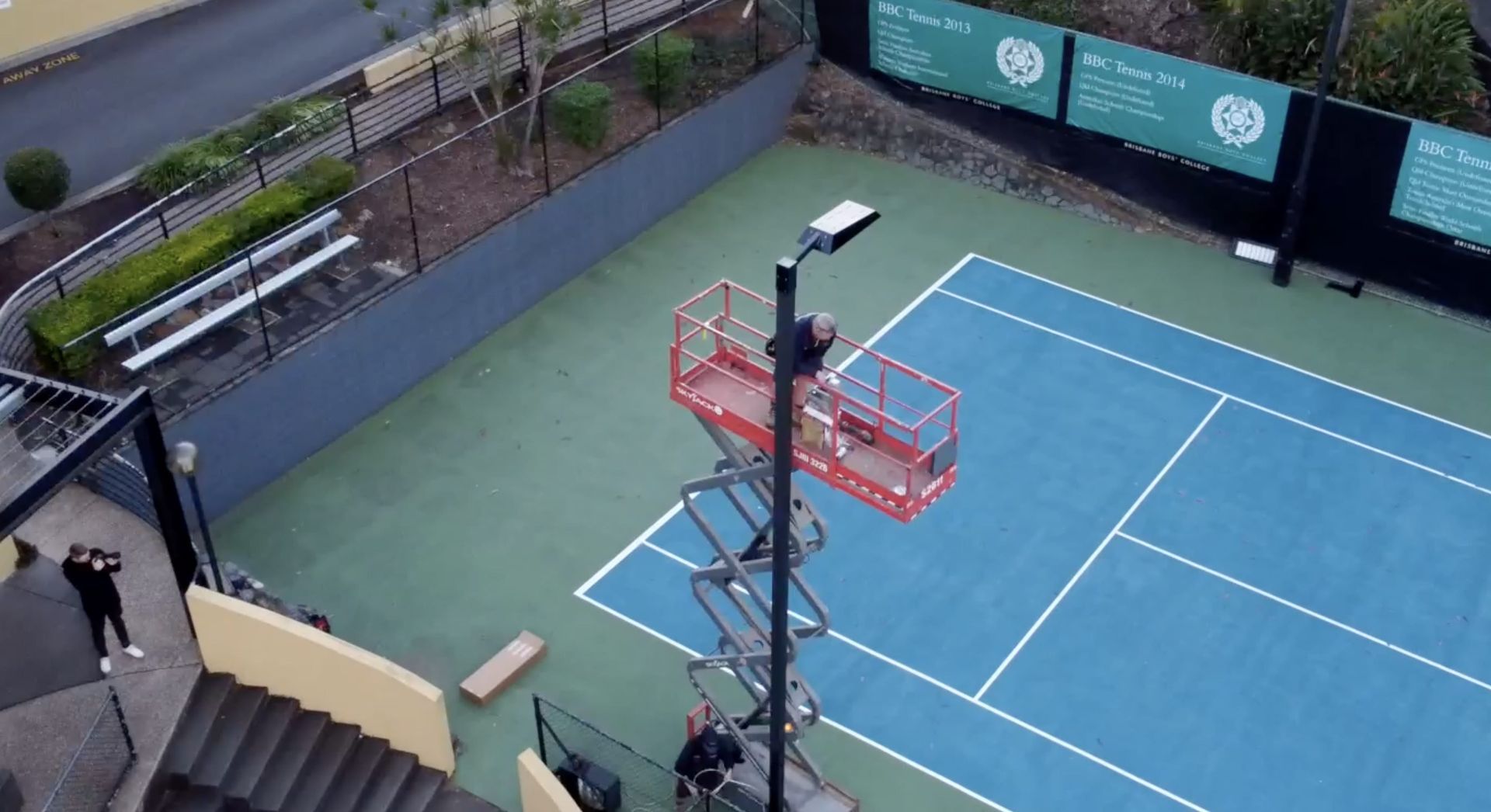 Overhead view of a blue tennis court with a tall light pole and a person walking by the stairs