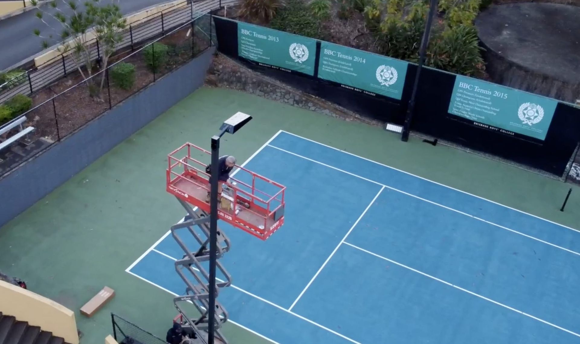 Empty blue tennis court with net, viewed from above beside teal sponsor banners and a tall light pole.