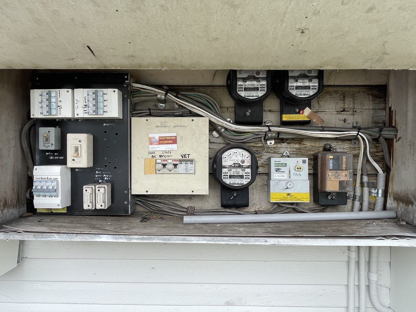 Electric utility meter and breaker panel array mounted in a wall recess with exposed wiring.