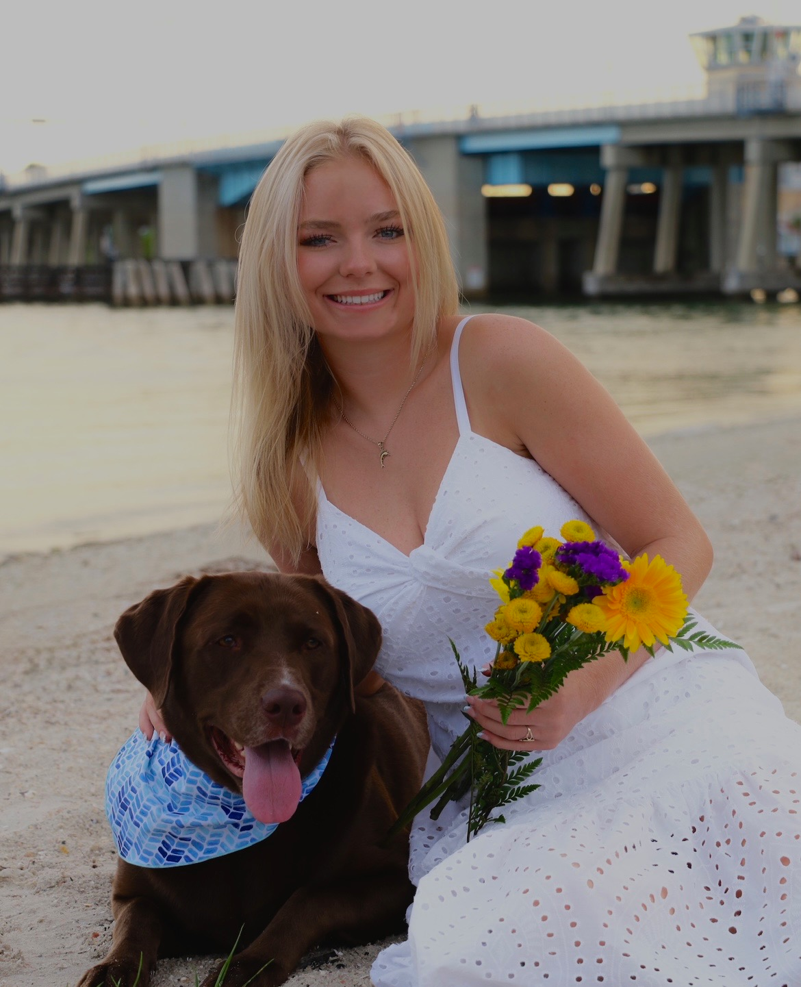 Woman in a white dress with a brown dog and bouquet by the water near a bridge