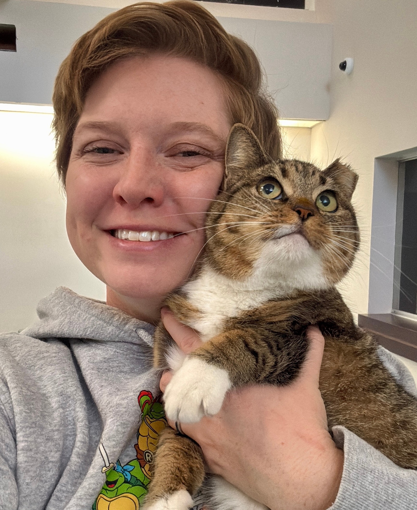 Woman in gray hoodie holding a tabby cat, both smiling. White paws, green eyes, indoor setting.