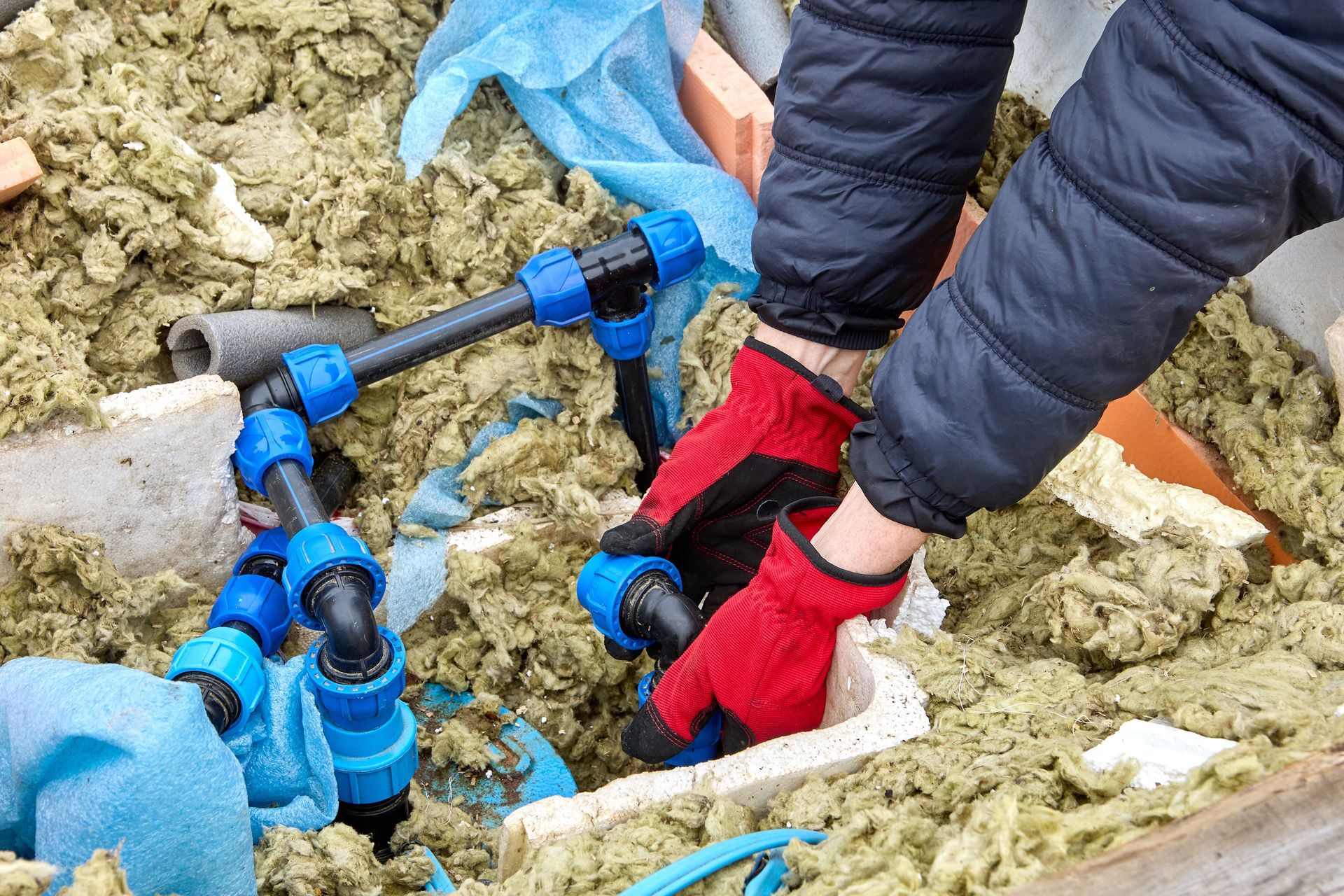 Gloved hands joining blue compression fittings on water pipes in insulation.