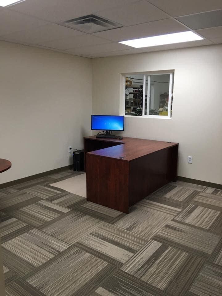 Empty office with a desk, computer monitor, and patterned carpet beneath fluorescent ceiling lights.