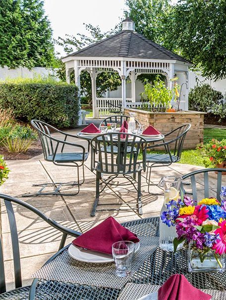 Patio dining area with metal table and chairs, red napkins, flowers, and a white gazebo in the background