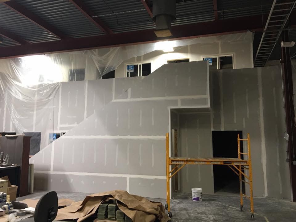 Partially finished interior wall in a warehouse, with drywall, scaffolding, and plastic sheeting.