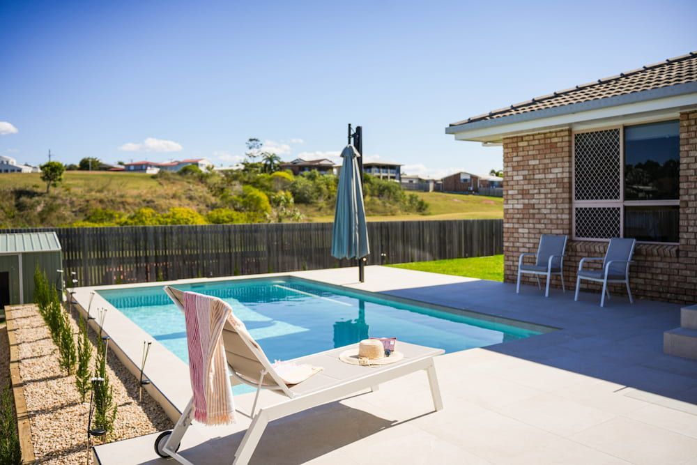 Backyard Pool With a Lounge Chair, Umbrella, and a Brick House — Concrete Pool Projects in Gladstone Central, QLD